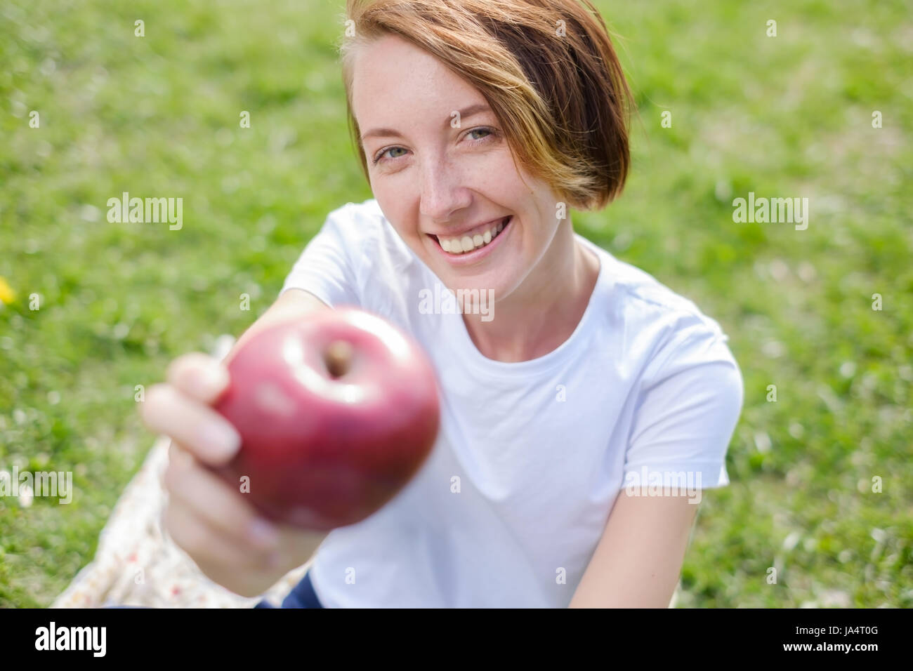 Molto bello il modello caucasico mangiare red apple nel parco. All'esterno ritratto di piuttosto giovane ragazza seduta su erba verde Foto Stock