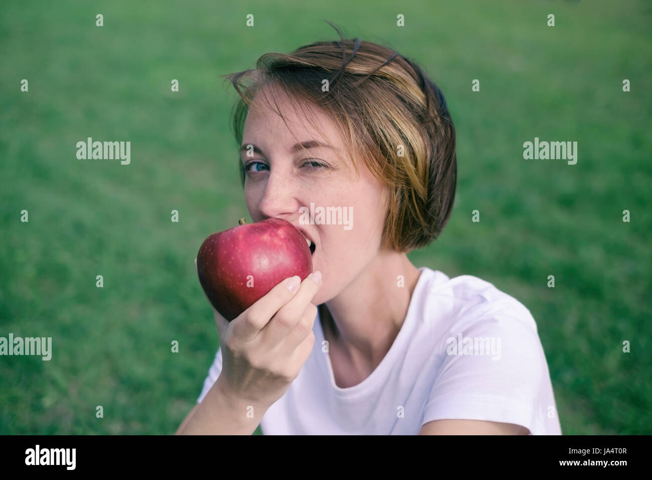 Molto bello il modello caucasico mangiare red apple nel parco. All'esterno ritratto di piuttosto giovane ragazza seduta su erba verde Foto Stock