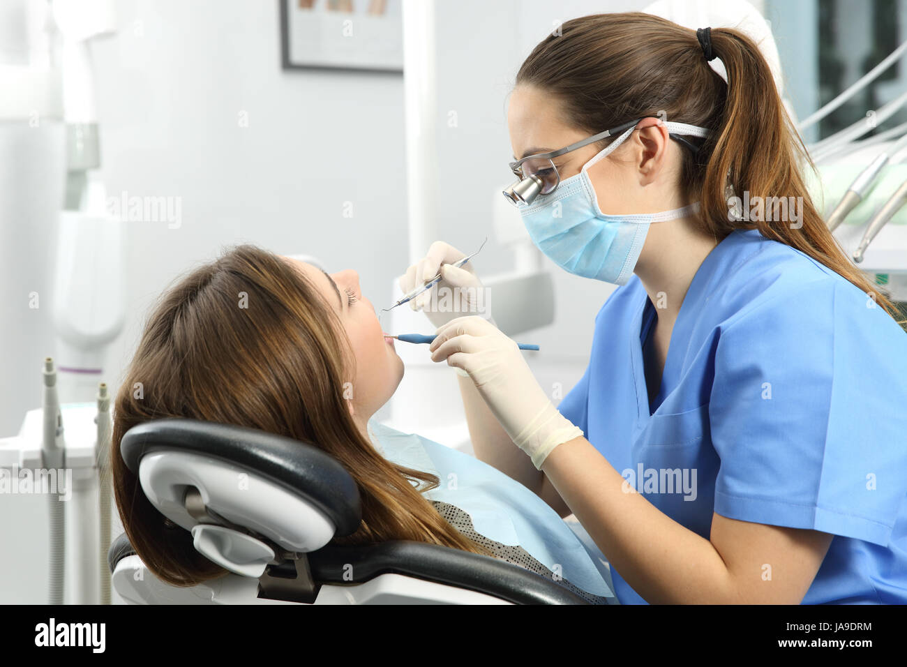 Dentista indossando occhiali guanti e maschera di esaminare un paziente denti con una sonda dentale e uno specchio in una clinica box con attrezzature in background Foto Stock