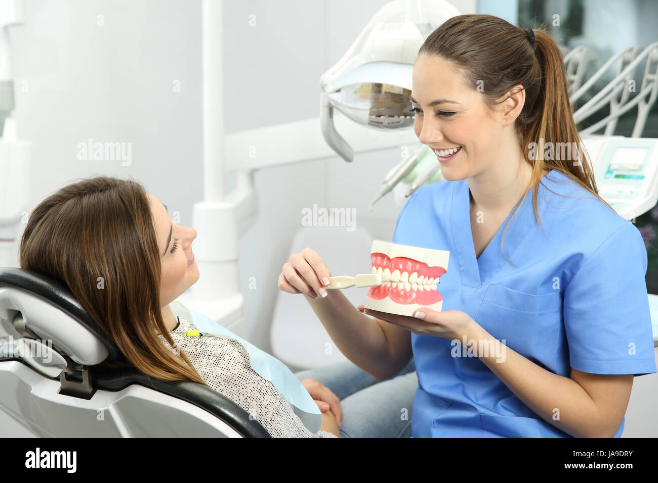 Dentista spiegando come spazzolare i denti correttamente per un paziente dopo i trattamenti seduto su una sedia in una clinica box con attrezzature mediche in backgro Foto Stock