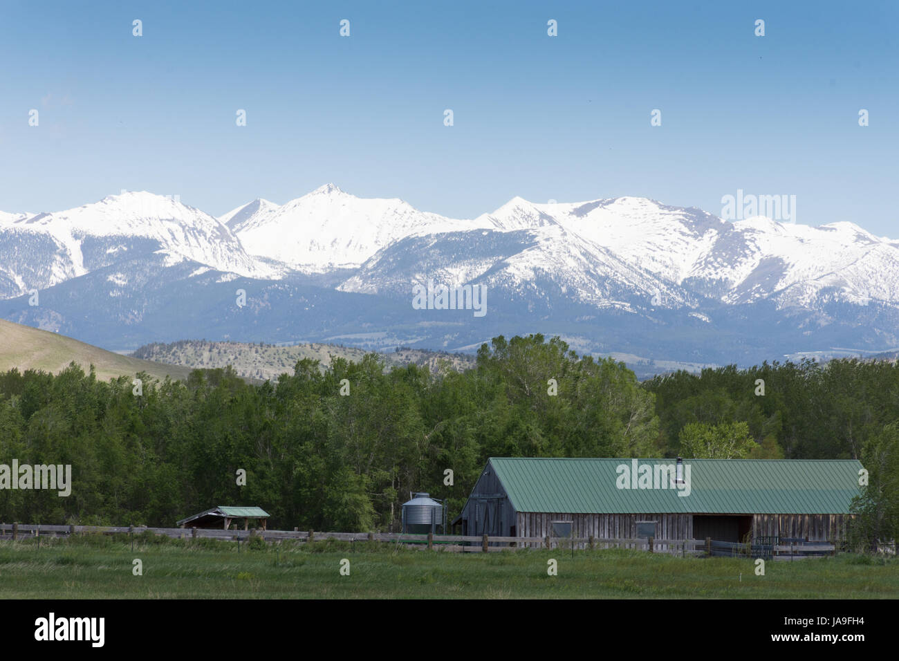 Fienile in campo verde con pascoli recintati in primo piano con aspre montagne innevate sullo sfondo. Foto Stock