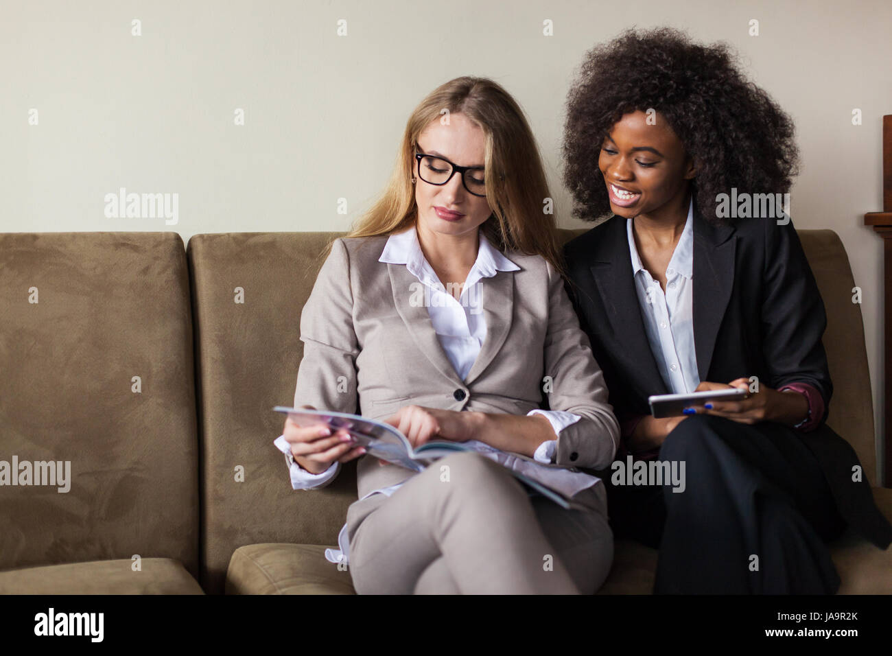 Due giovani e belle imprenditrici seduto sul divano con la compressa e la  rivista nelle loro mani. Una donna è nero con capelli ricci. Donne che  indossano tute Foto stock - Alamy