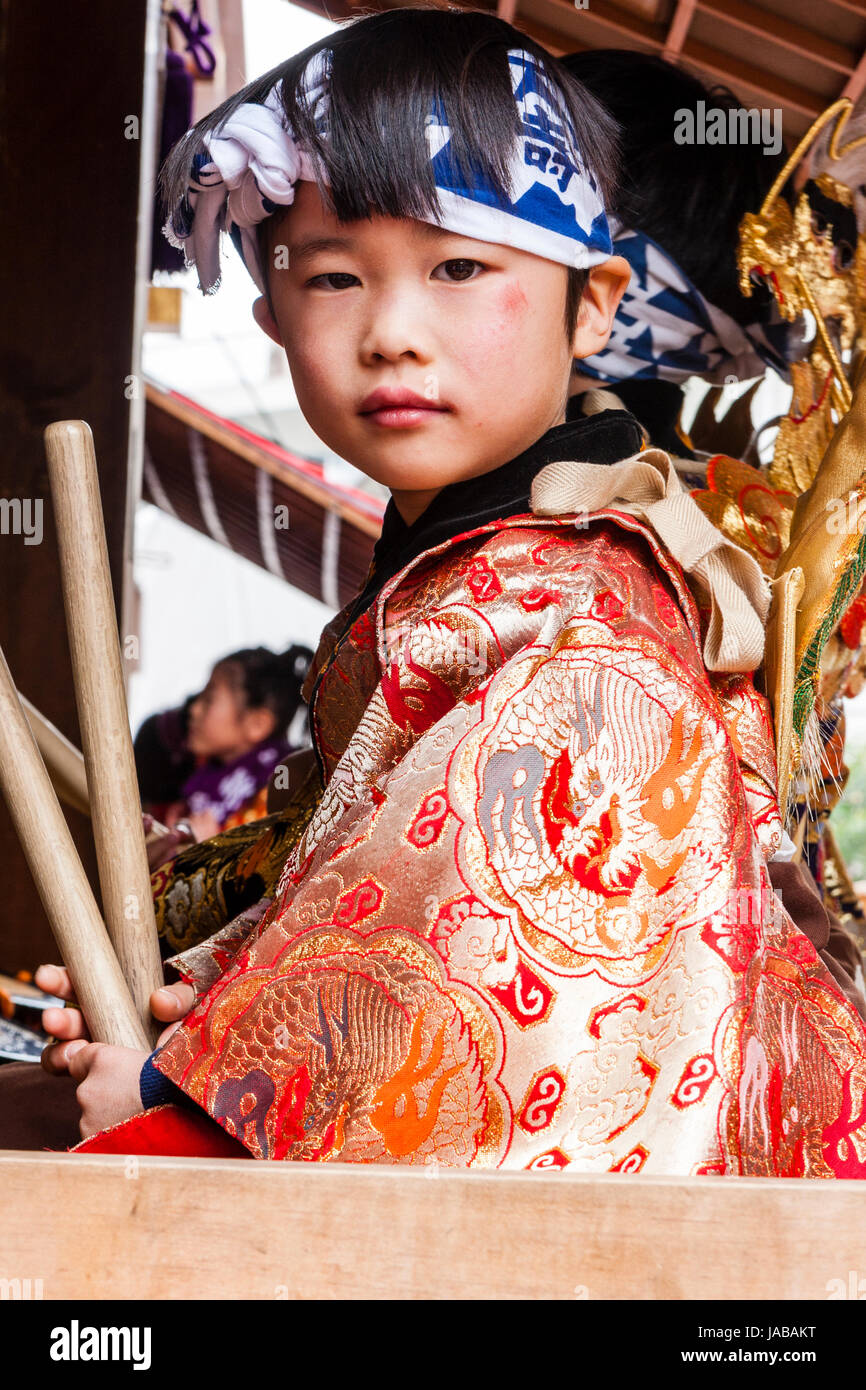 Asian, bambino giapponese, ragazzo, molto viso carino, in seduta Dashi galleggiante tenendo un paio di bastoncini di tamburo. Indossa il kimono arancione e la fascia di testa. Contatto visivo. Foto Stock