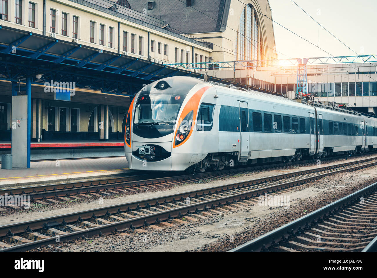 Treno ad alta velocità presso la stazione ferroviaria al tramonto in Europa. Moderno treno intercity sulla linea ferroviaria piattaforma. Paesaggio industriale con treni passeggeri Foto Stock