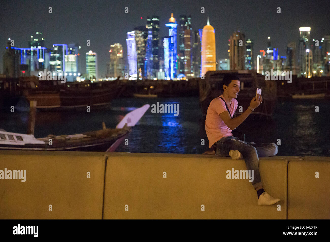 Un giovane uomo si siede sulla parete del mare di Corniche, effettuare una video chiamata usando il suo telefono cellulare. Sullo sfondo il Dhow Harbour e al di là della hi Foto Stock