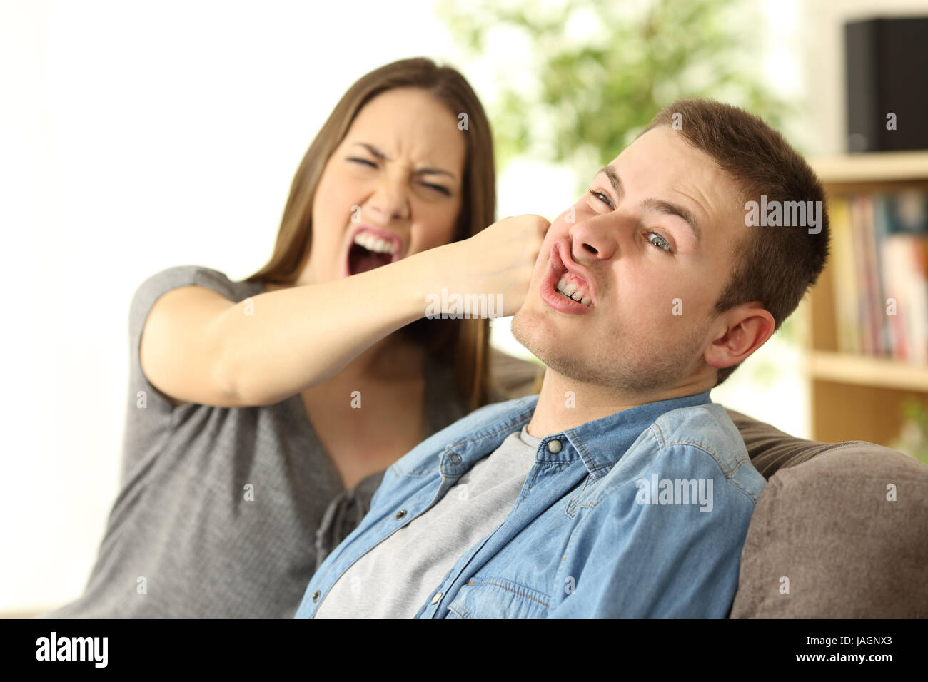 Donna di colpire il suo ragazzo seduto su un divano nel soggiorno in un interno di una casa Foto Stock