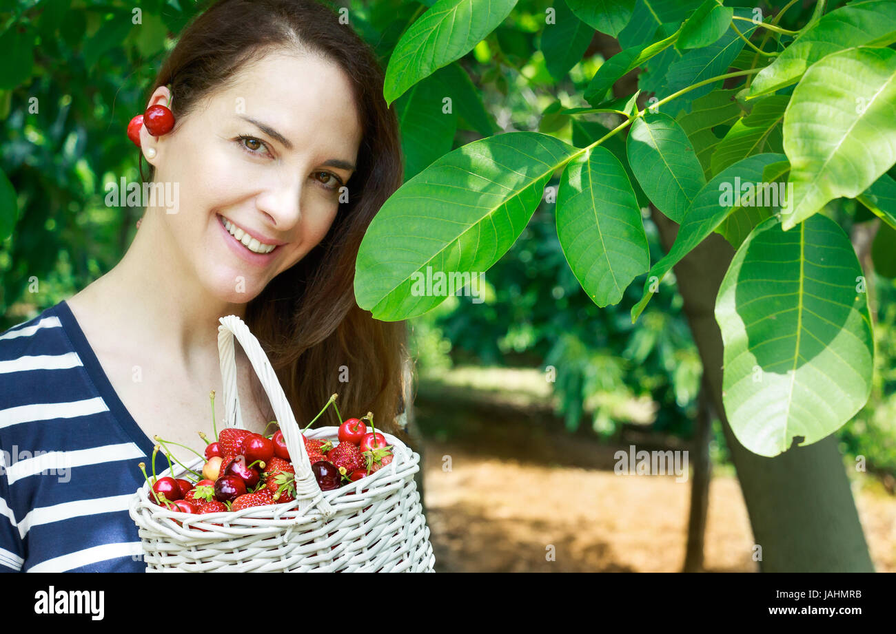 Una metà di età della donna rappresenta un cesto pieno di ciliegie, sorridente, guardando la fotocamera. Foto Stock