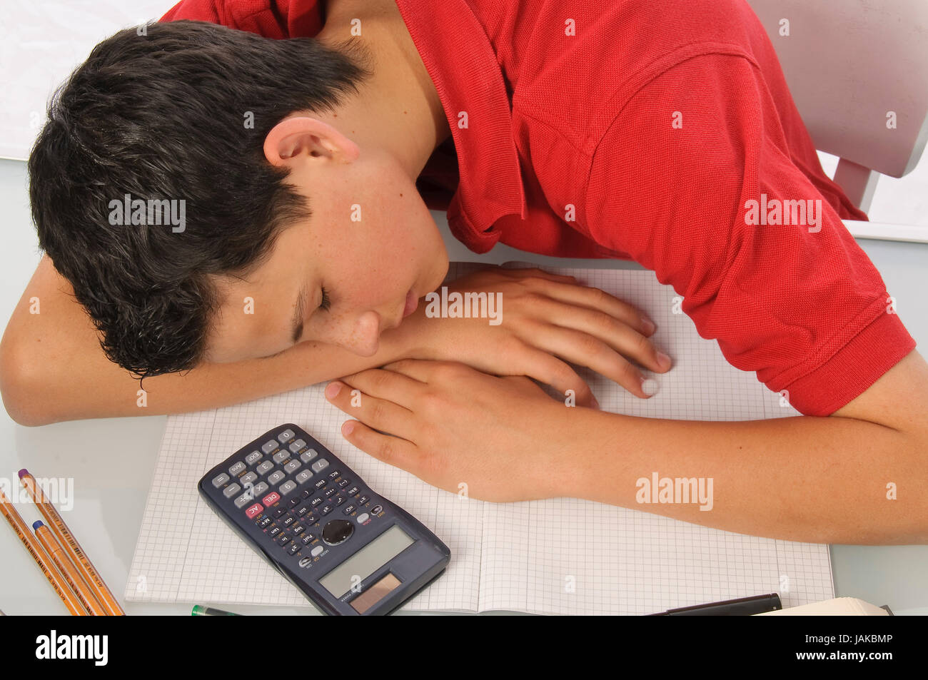 Junge erschöpft sitzt und frustriert über Hausaufgaben am Schreibtisch vor weißem Hintergrund Foto Stock