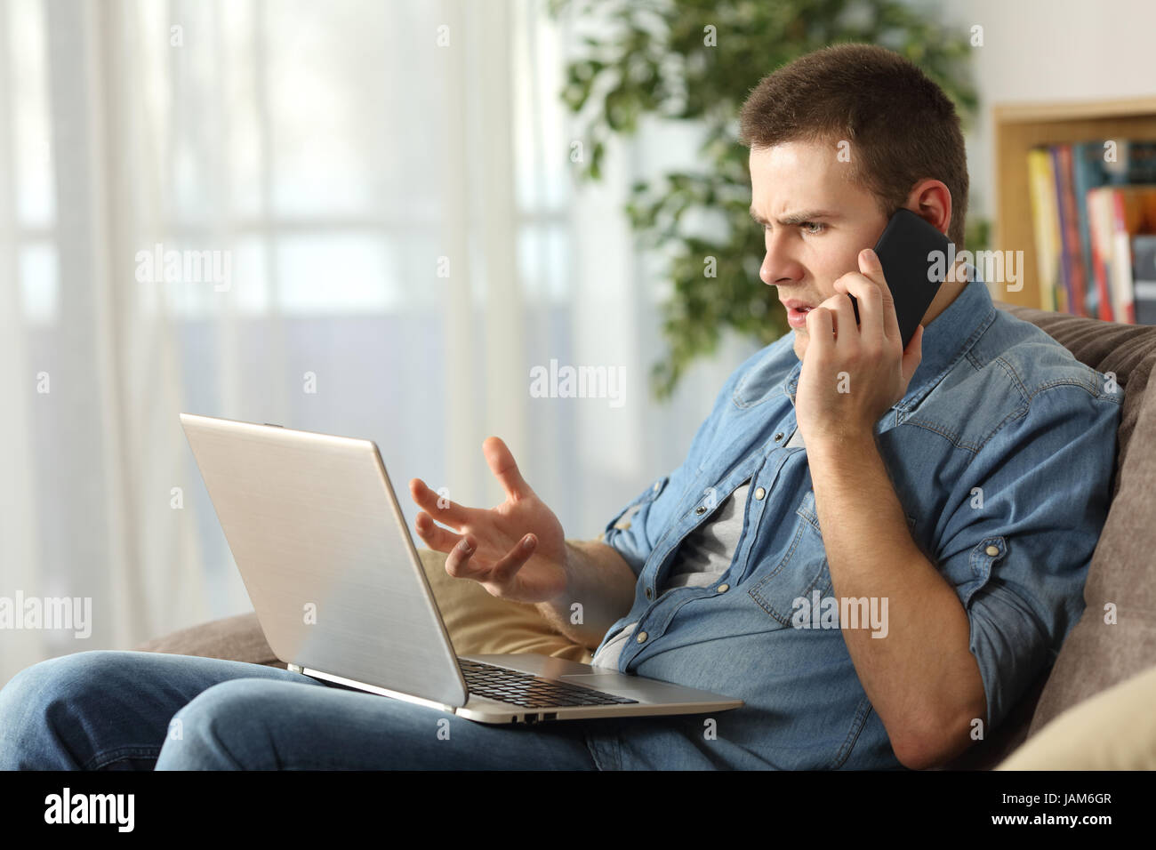 Angry Man aventi problemi sulla linea con un laptop a parlare al telefono cellulare con il servizio di supporto seduta su un divano nel salotto di casa Foto Stock
