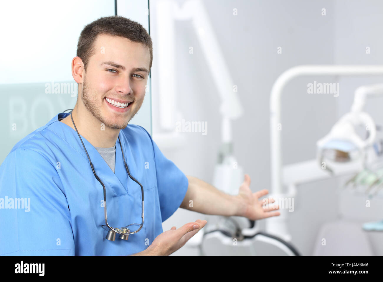 Medico indossando blue coat vi invita al suo studio dentistico e guardando la fotocamera con attrezzature mediche in background Foto Stock