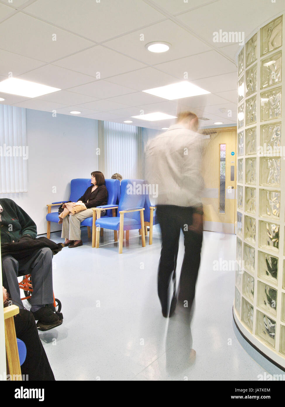 Seduta di pazienti in ospedale NHS area di attesa, come medico passeggiate attraverso il telaio, offuscata attraverso il movimento Foto Stock