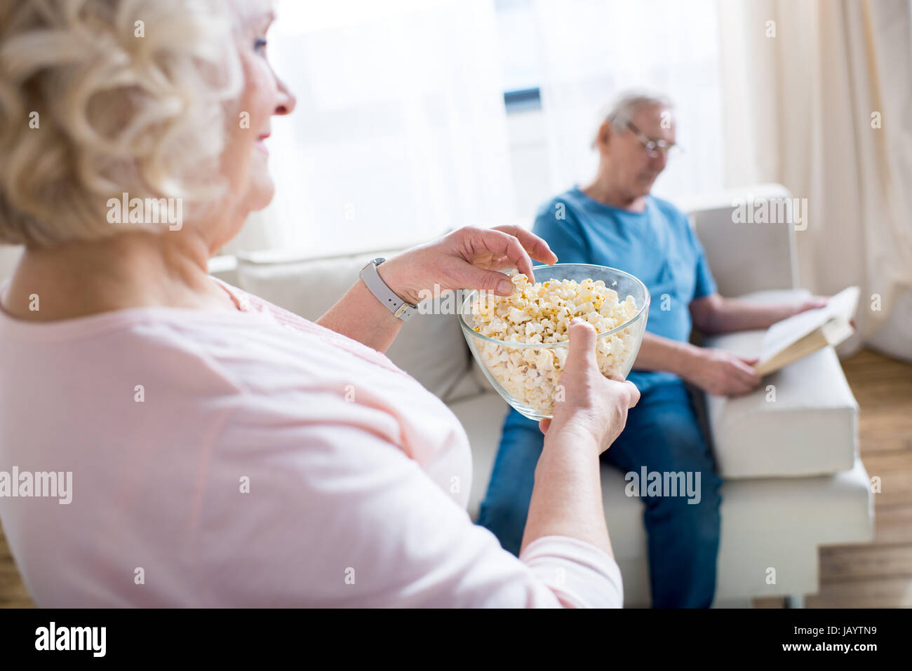 Senior donna mangiare popcorn, uomo seduto sul divano e la lettura prenota dietro Foto Stock