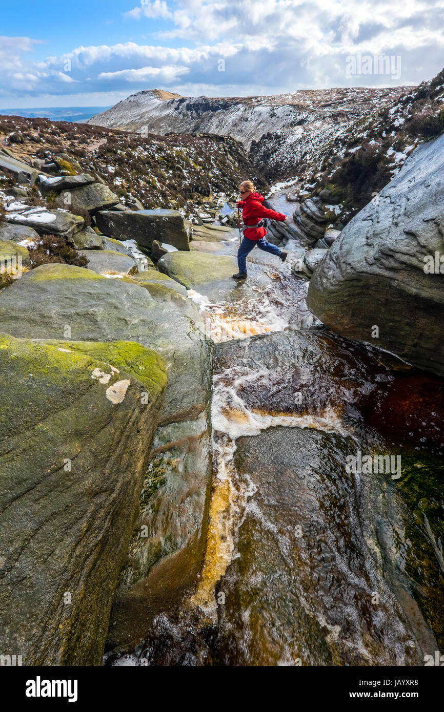 Una femmina di walker attraversare un torrente in inverno in cima Grindsbrook sul bordo meridionale di Kinder Scout, Parco Nazionale di Peak District Foto Stock