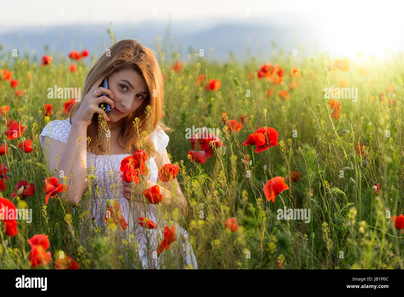 Ragazza giovane indossando abito bianco panno in piedi in un campo di papavero con il suo telefono Foto Stock