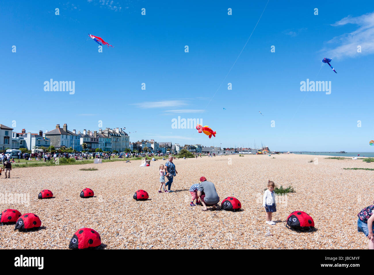 Ladybird tipo aquiloni fissato alla spiaggia sassosa mentre altri aquiloni galleggiante nel cielo azzurro. Ampio angolo dell'immagine. Trattativa, Inghilterra, il lungomare come sfondo. Aquiloni evento. Foto Stock