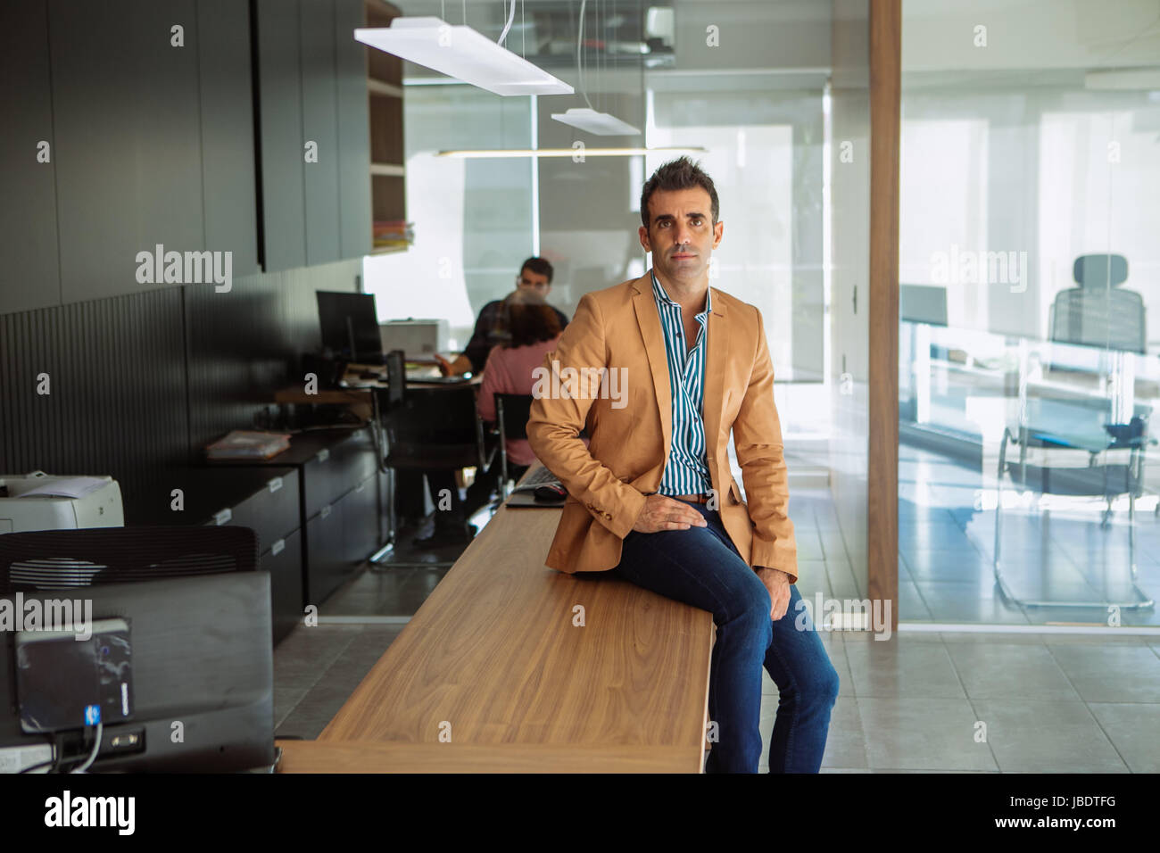 Un 35 - 40 anni di vecchio uomo caucasico capelli scuri cool look moderno in un ambiente informale, maglietta, blazer e jeans di posti a sedere su una scrivania in un ufficio Foto Stock