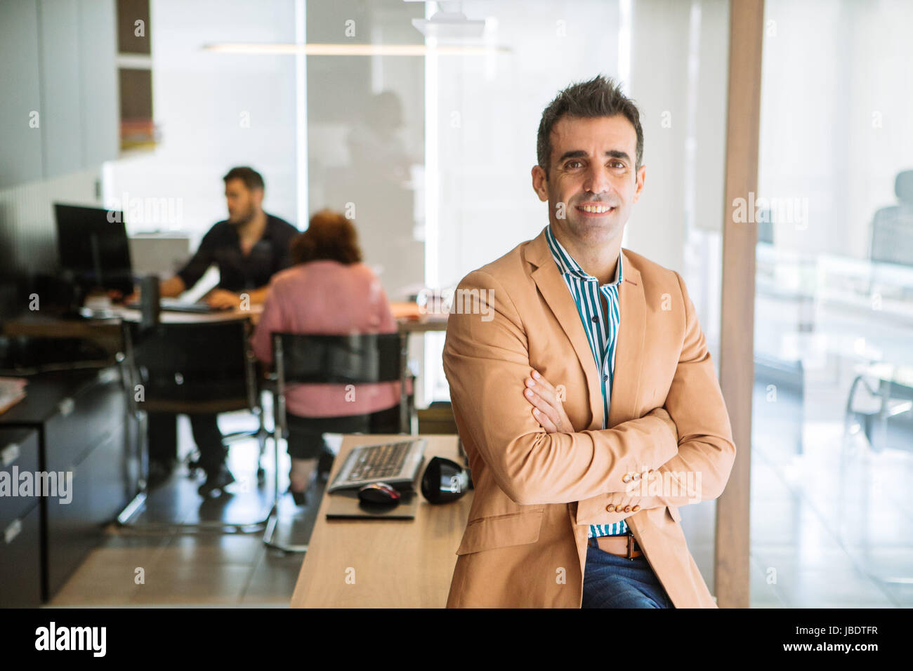 Un 35 - 40 anni di vecchio uomo caucasico capelli scuri cool look moderno bracci ripiegati sorridente in un spogliato della camicia, blazer e jeans di posti a sedere su una scrivania in un ufficio Foto Stock