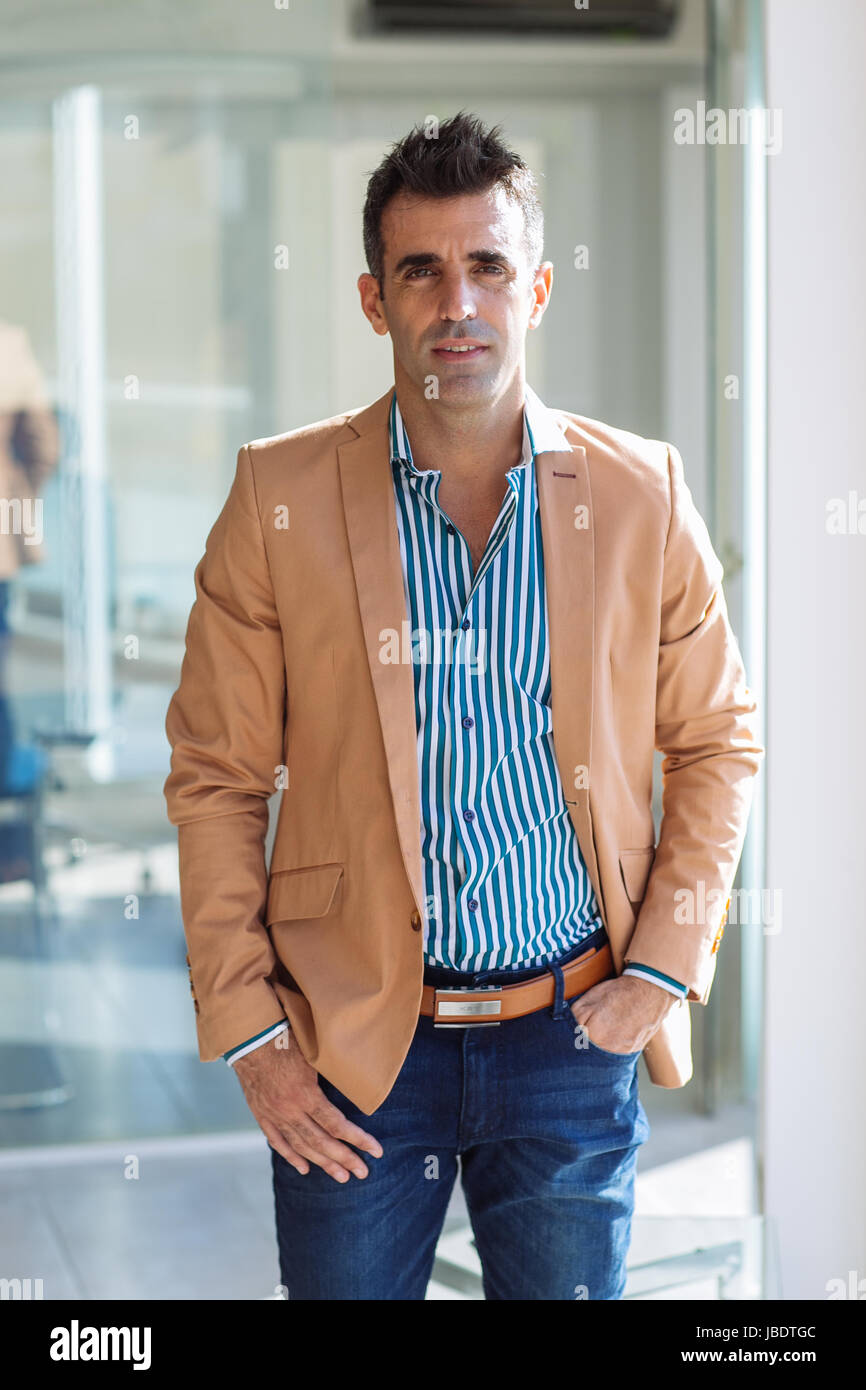 Un 35 - 40 anni di vecchio uomo caucasico capelli scuri cool look moderno con mano nella tasca di una camicia spellata, blazer e jeans in un ufficio di medie colpo lungo Foto Stock