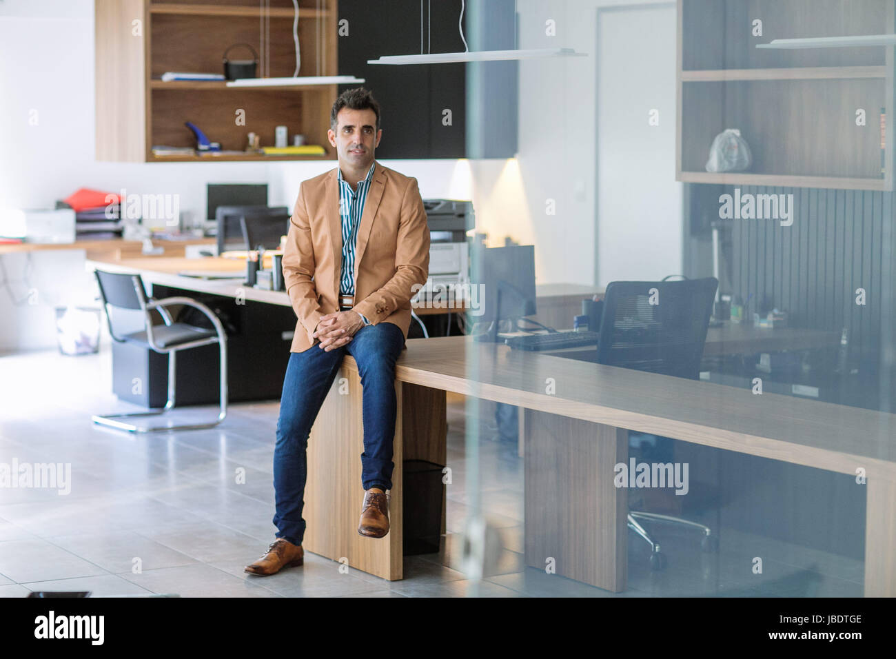Un 35 - 40 anni di vecchio uomo caucasico capelli scuri cool look moderno sorridente in un spogliato della camicia, blazer e jeans di posti a sedere su una scrivania in un ufficio vuoto Foto Stock
