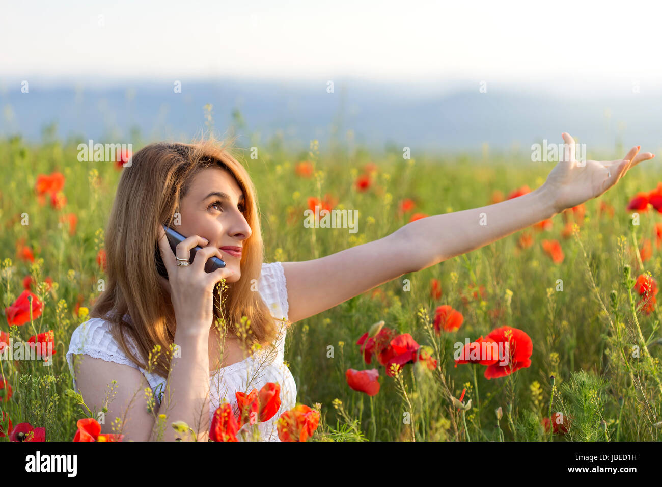 Ragazza giovane indossando abito bianco panno in piedi in un campo di papavero parlando al telefono Foto Stock