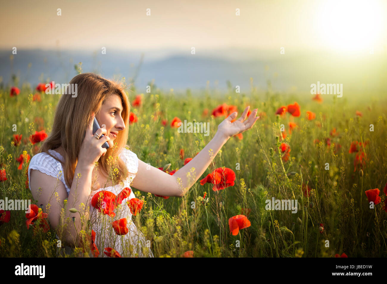 Ragazza giovane indossando abito bianco panno in piedi in un campo di papavero parlando al telefono Foto Stock