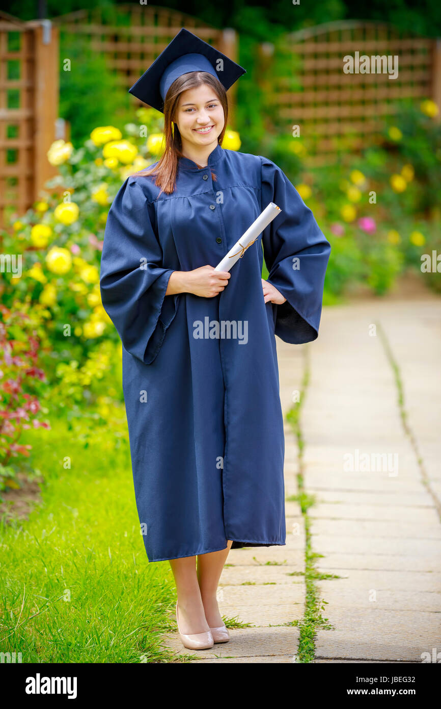 Sorridente giovane donna diploma di contenimento e indossando il cappello e camice all'esterno guardando la fotocamera. Concetto di graduazione. Foto Stock