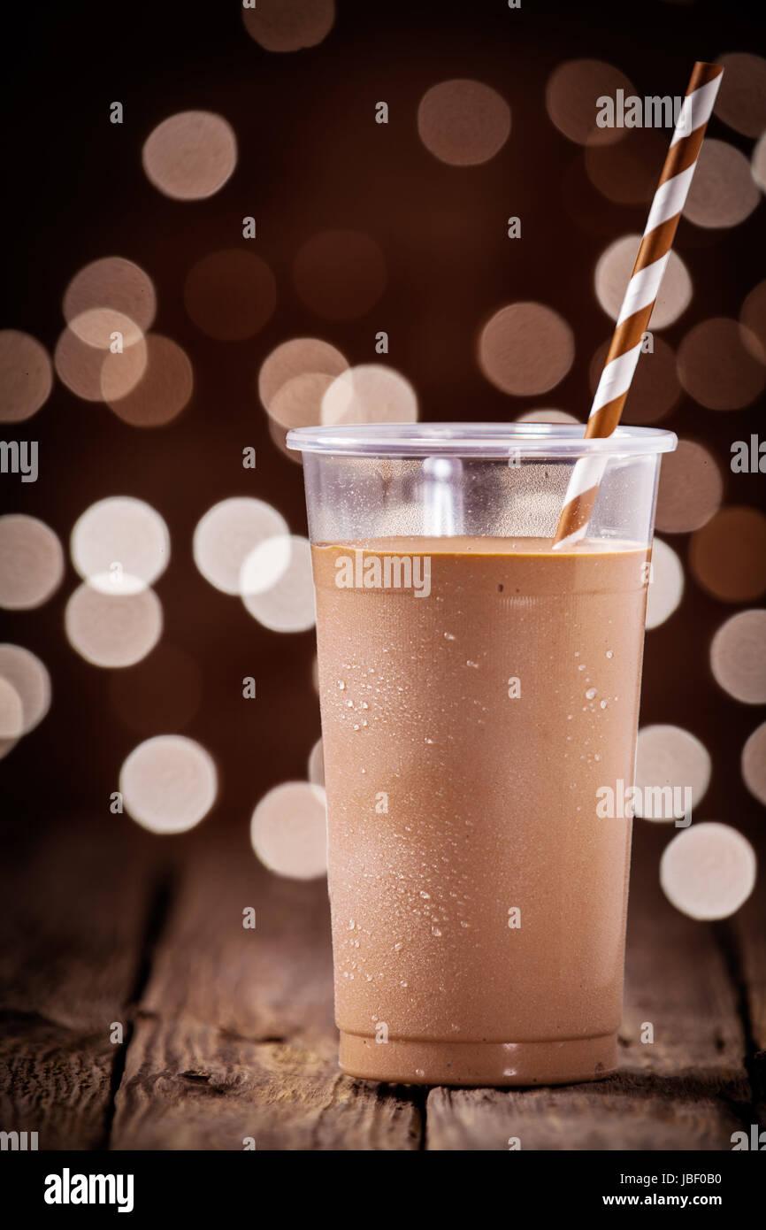 Deliziosa refrigerate da asporto frullato al cioccolato in un bicchiere di plastica in piedi su una tavola in legno rustico contro uno sfondo bokeh di scintillanti luci festive Foto Stock