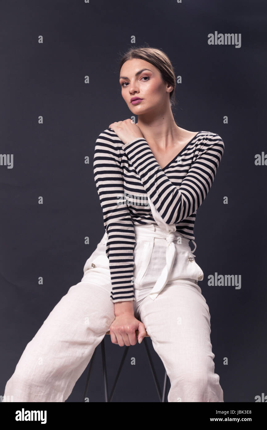 Una giovane donna caucasica 20s, 20-29 anni, moda modello seduta sgabello da bar, ponendo, studio, sfondo bianco, striscia orizzontale t-shirt, pantaloni bianchi, l Foto Stock
