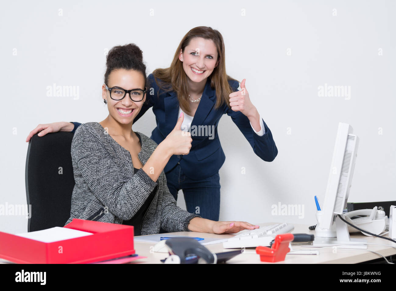Zwei Frauen am Schreibtisch im Büro zeigen mit dem Daumen nach oben und lächeln in die Kamera. Foto Stock