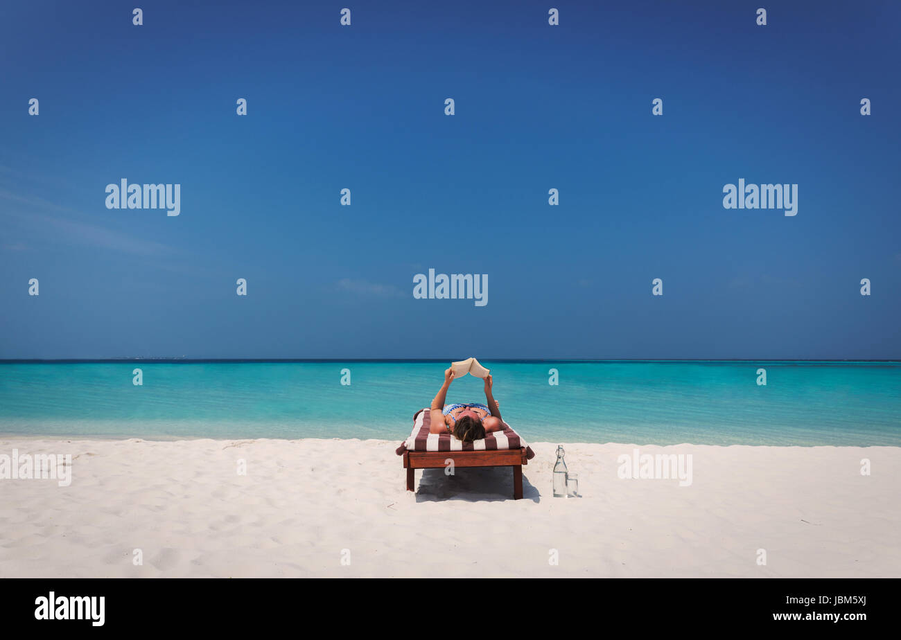 Donna rilassante, posa in opera e la lettura di libro sulla sedia a sdraio sulla soleggiata spiaggia tropicale Foto Stock