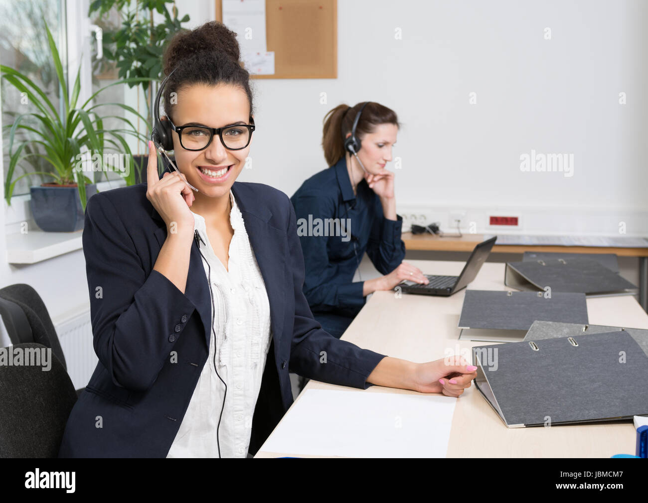Eine junge Büroangestellte mit Headset sitzt vor ihren Akten im Büro. Eine weitere Frau mit Headset befindet sich im Hintergrund (unscharf). Foto Stock