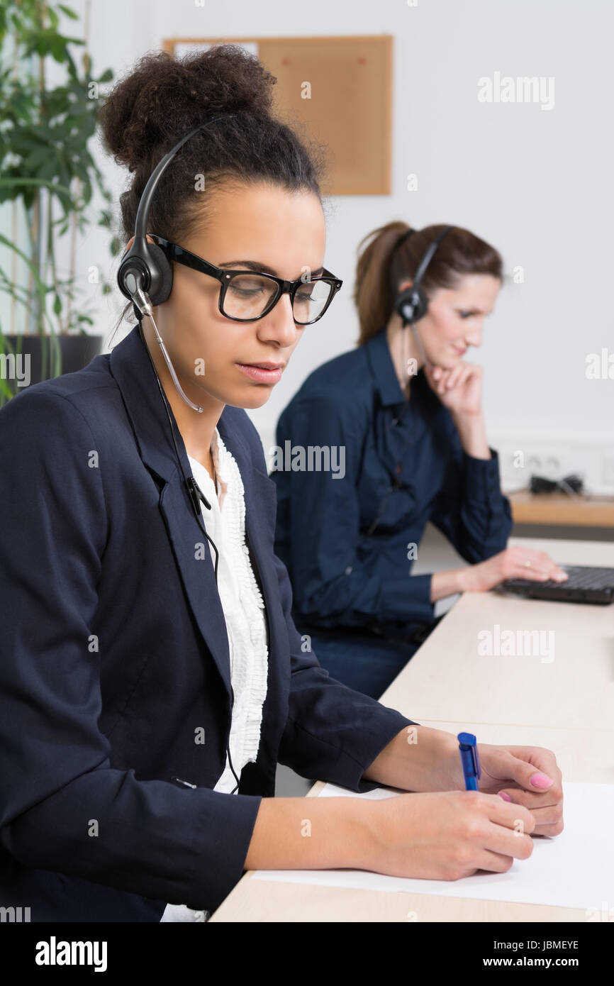 Eine junge Büroangestellte mit Headset schreibt in ein Dokument. Eine weitere Frau mit Headset befindet sich im Hintergrund (unscharf) vor einem Notebook. Foto Stock