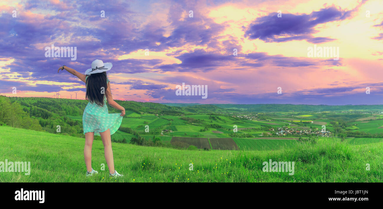 Panoramica estiva con un europeo brunette donna vestito con un abito verde con un cappello bianco sul suo capo, godendo del tramonto su una collina Foto Stock