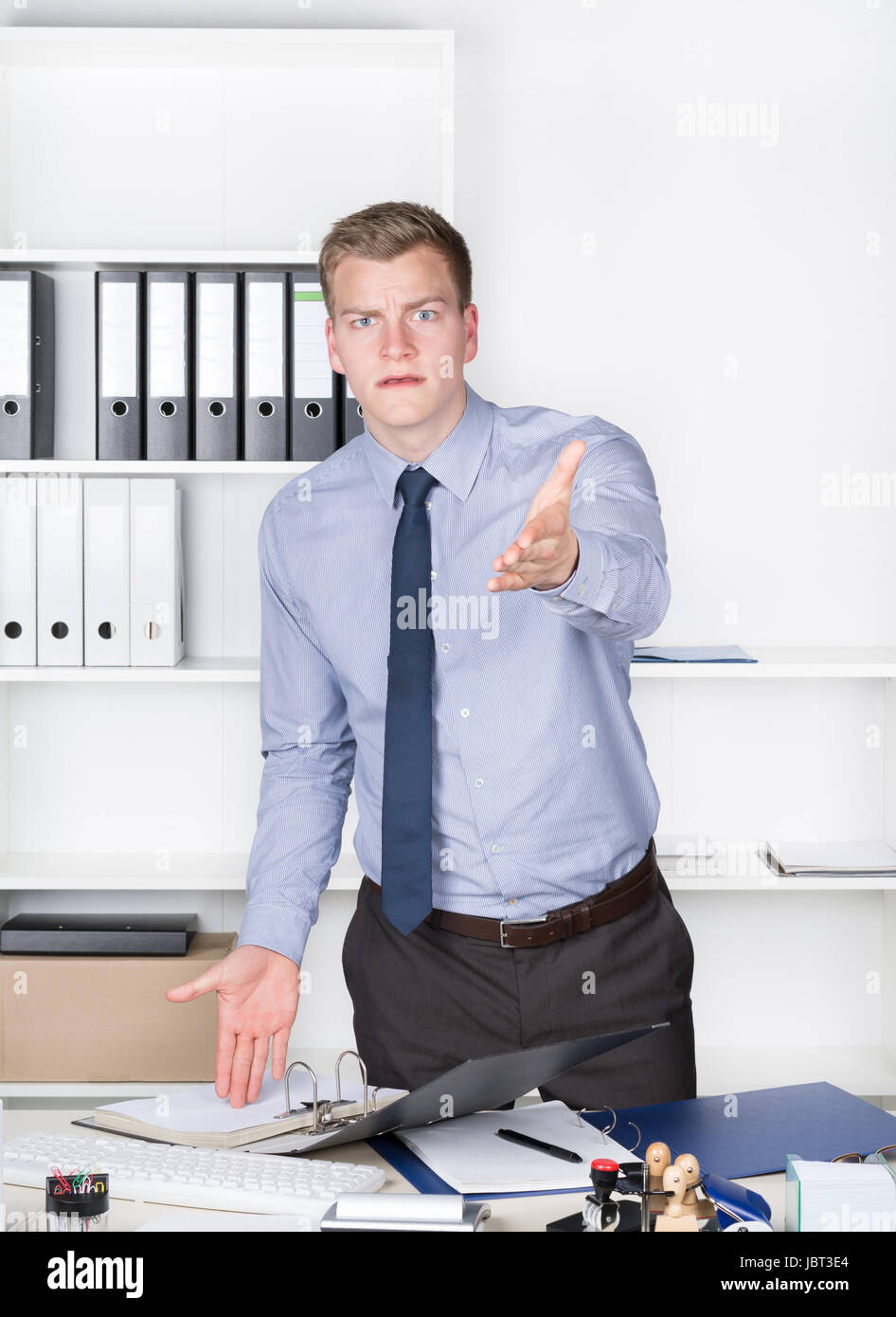 Junger wütender Geschäftsmann steht vor einem Regal im Büro und streckt einem braccio aus in Richtung Kamera. Der Mann schaut zur Kamera. Foto Stock