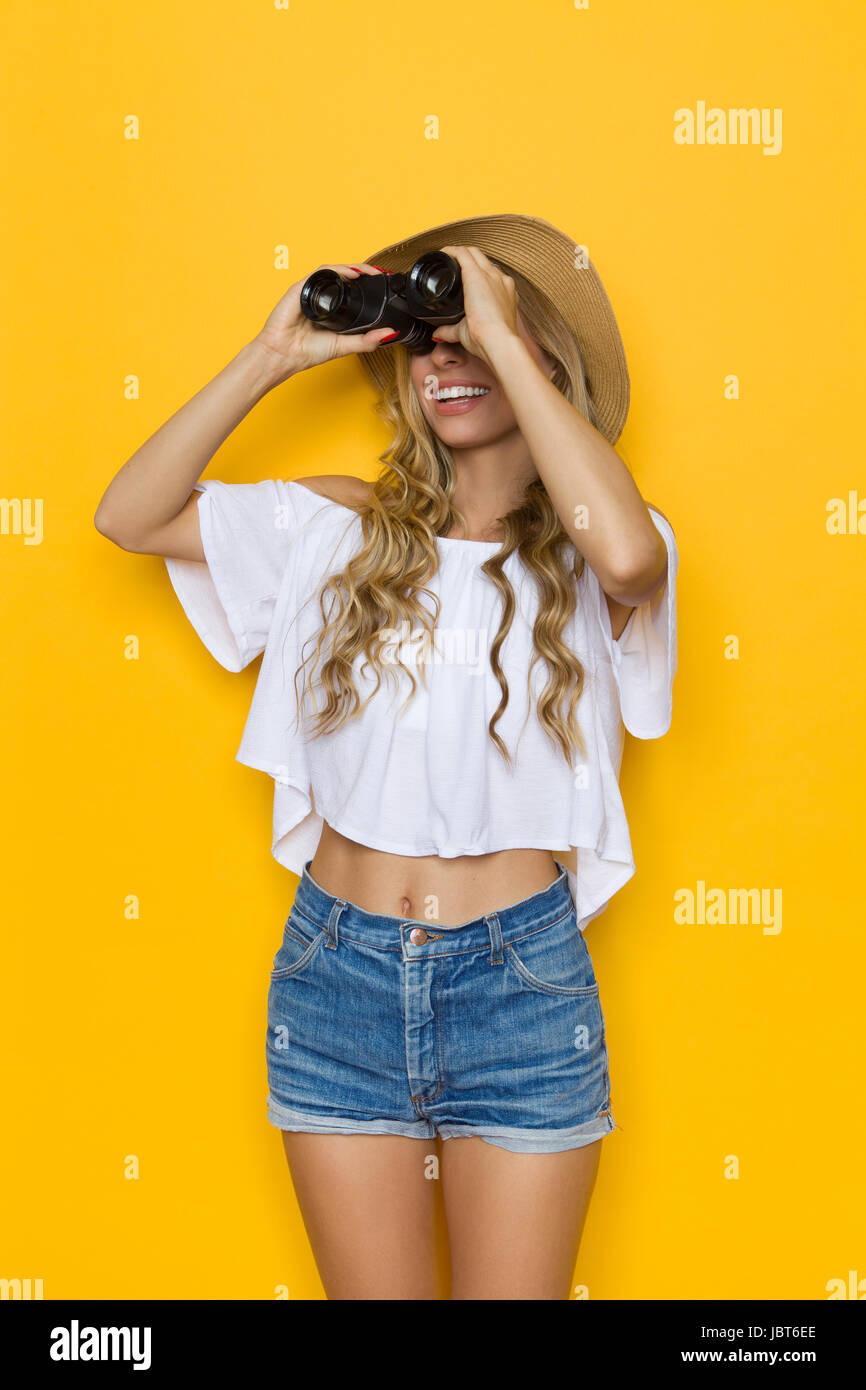 Giovane donna bionda in jeans corti, una camicia bianca e cappello di  paglia, guardando lontano attraverso il binocolo, tre quarti di lunghezza  studio shot su sfondo giallo Foto stock - Alamy