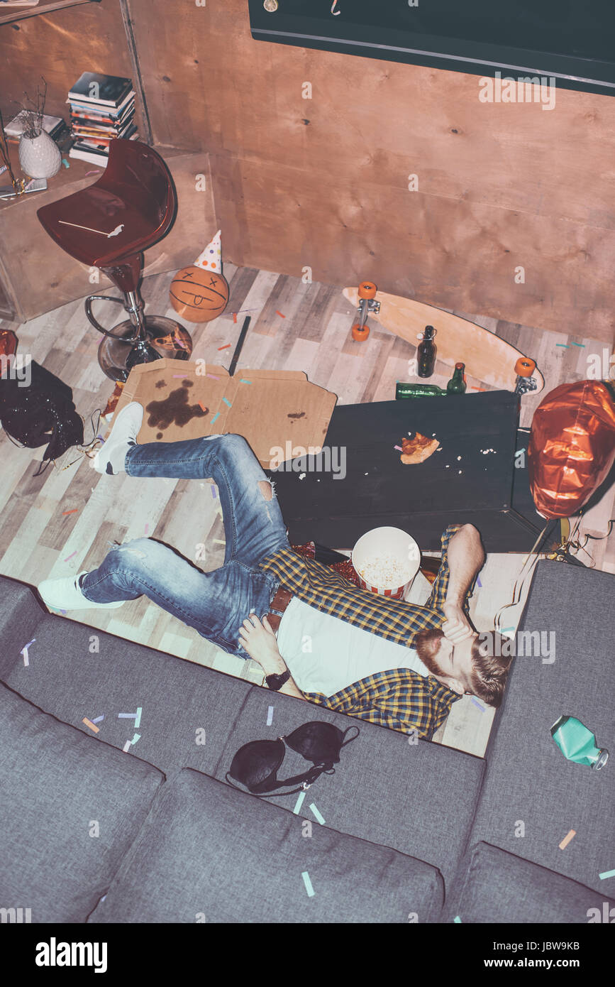 Ubriaco uomo barbuto sdraiato sul pavimento in camera disordinato after party Foto Stock
