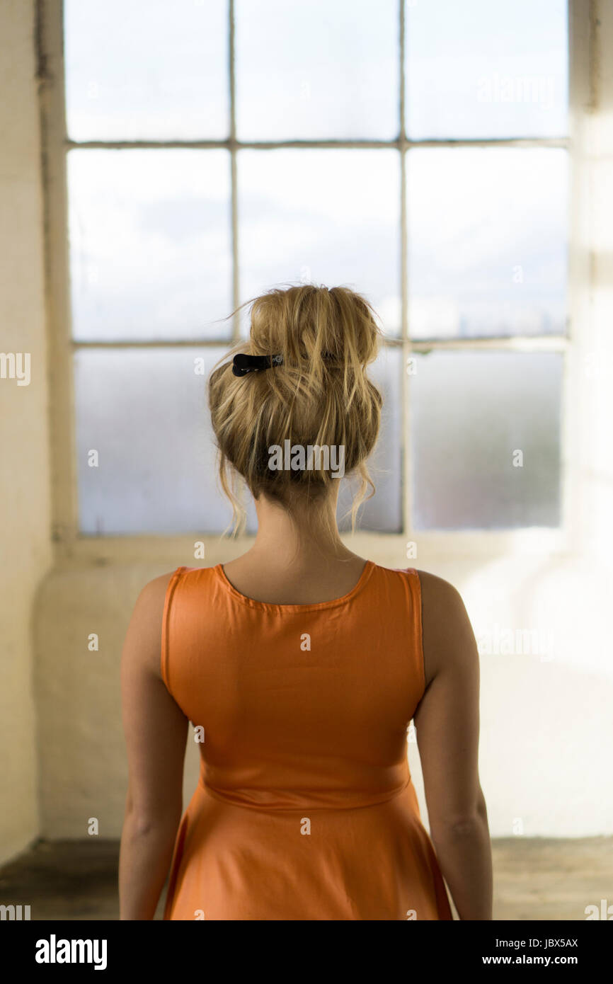 Vista posteriore di un giovane biondo woamn caucasica indossando un arancio abiti estivi in piedi dalla finestra Foto Stock