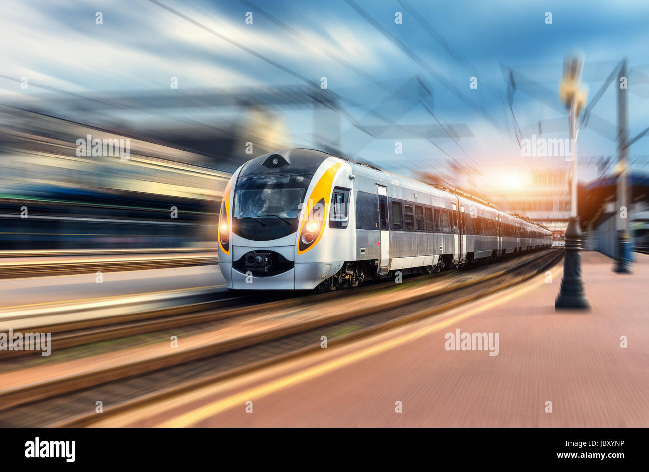 Treno ad alta velocità in movimento con la stazione ferroviaria al tramonto. Europeo moderno treno intercity sulla piattaforma ferroviaria con motion blur effetto. Industri Foto Stock