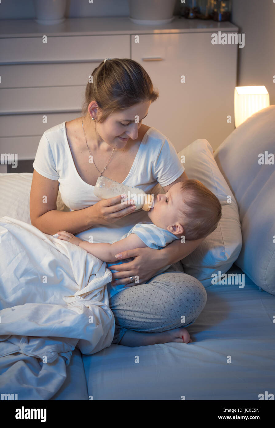 Ritratto di giovane madre premurosa dando una bottiglia con latte per il suo bambino a letto a notte. Concetto di Parenting Foto Stock