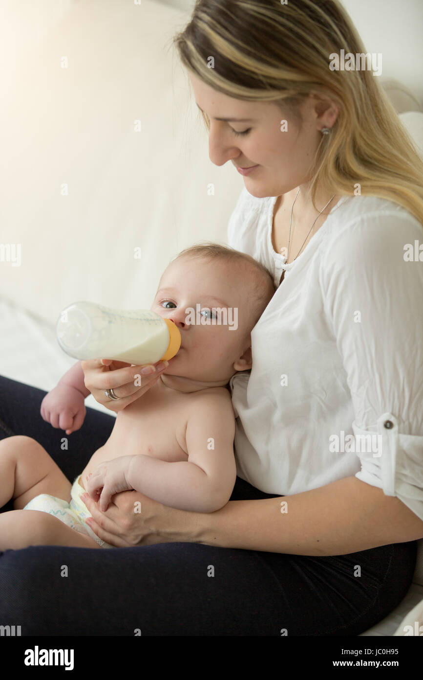 Tonica Ritratto di giovane madre seduta sul letto dando bottiglia al suo bambino Foto Stock