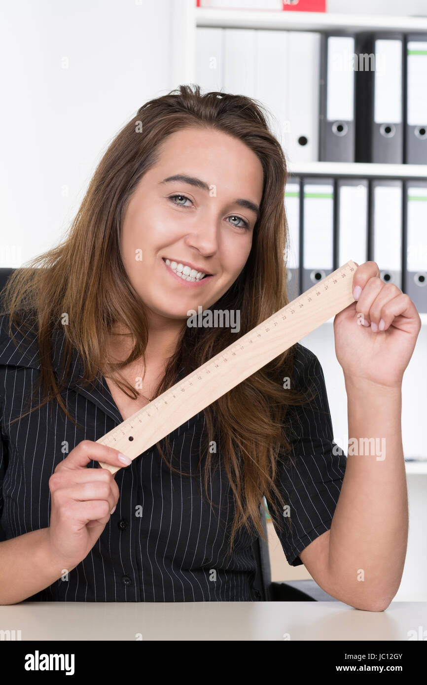Eine junge, lächelnde Frau hält ein Lineal aus Holz am Schreibtisch im Büro. Im Hintergrund steht ein regale. Foto Stock