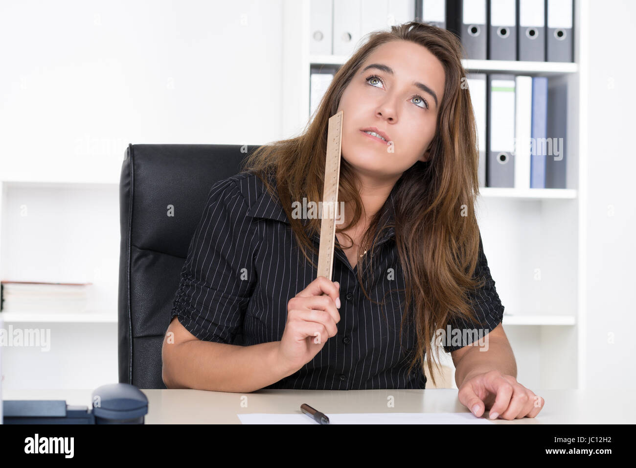 Eine junge, nachdenkliche Frau hält ein Lineal aus Holz un ihre Wange während sie am Schreibtisch im Büro sitzt. Im Hintergrund steht ein regale. Foto Stock