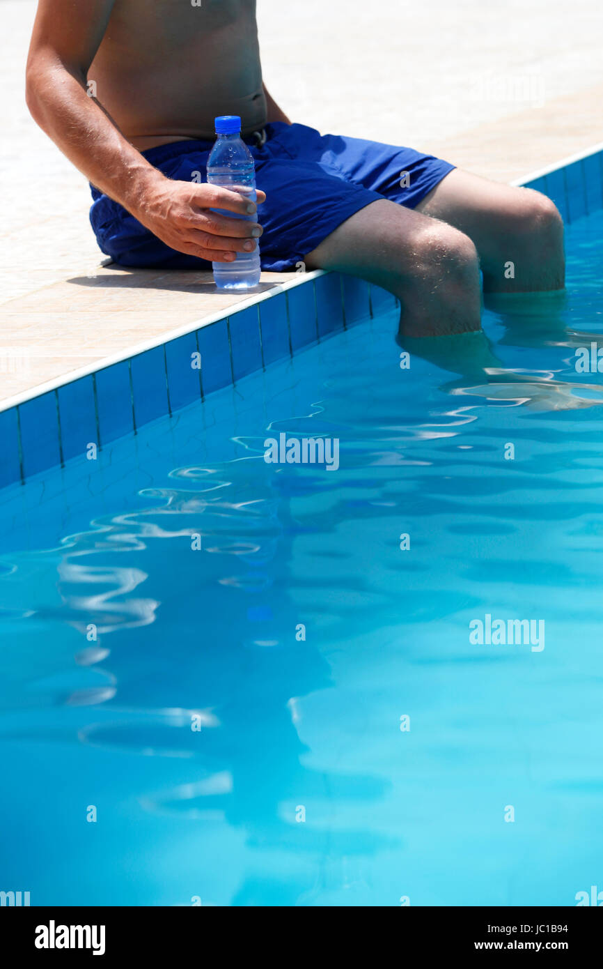 Attraente uomo con costume da bagno blu e una bottiglia di acqua gode di sole in una piscina Foto Stock