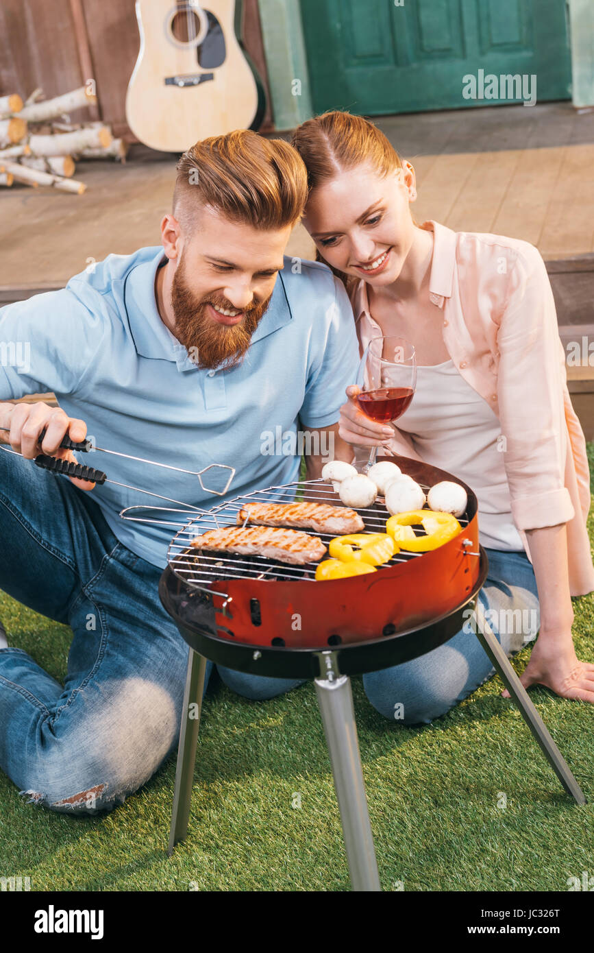 L uomo e la donna la tostatura la carne e le verdure su barbecue grill, donna holding wineglass Foto Stock
