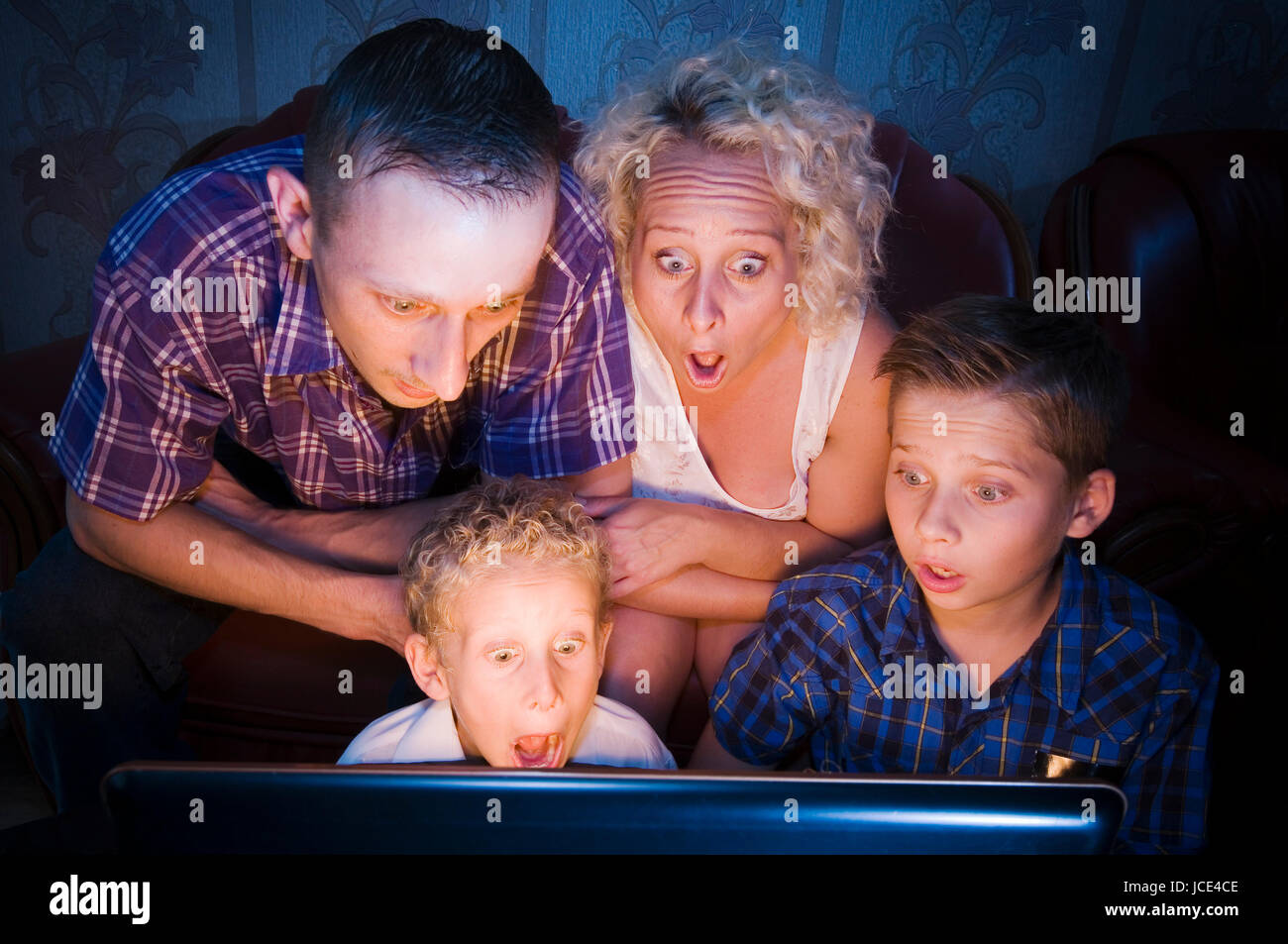 Eltern mit ihren zwei Söhnen im Alter von 7 und 13 Jahren abgedunkelten im Zimmer auf der Couch sitzend und schockiert in einen Monitor in Richtung Kamera blickend Foto Stock