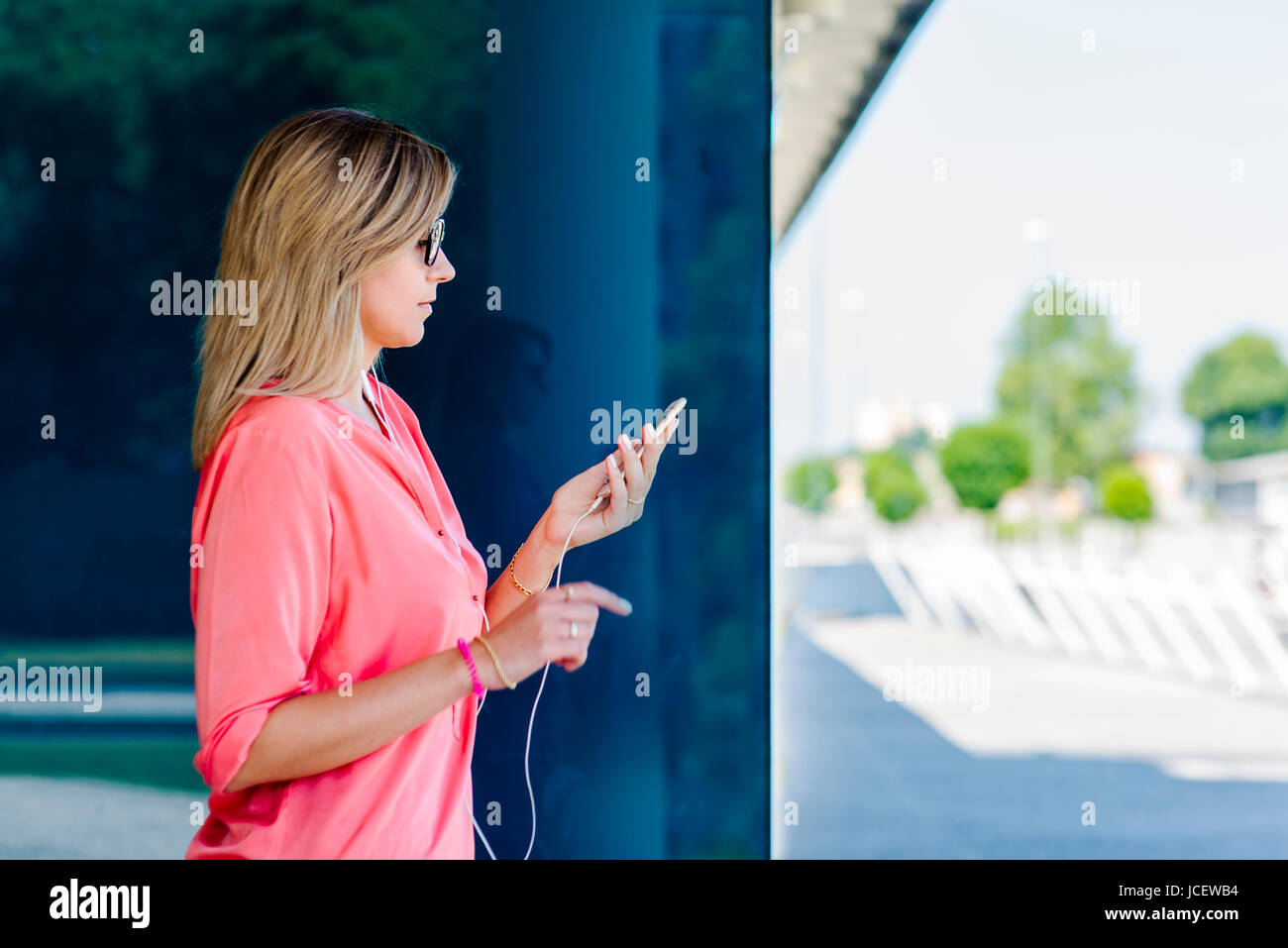 Ritratto di una giovane donna caucasica fiduciosa e curvilinea in un quartiere moderno della grande città con smartphone, auricolari, connessi, all'aperto, luce del giorno. Foto Stock