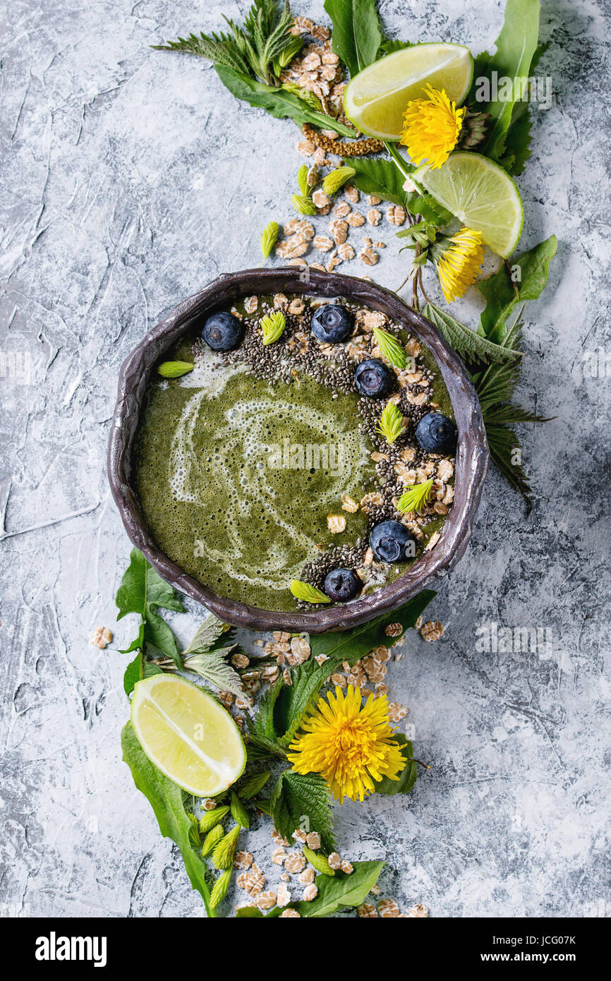 Verde primavera ortica e tarassaco ciotola smoothie servita con calce, fiori gialli, foglie giovani, fiocchi di avena, chia semi, mirtilli, crema su grigio b Foto Stock