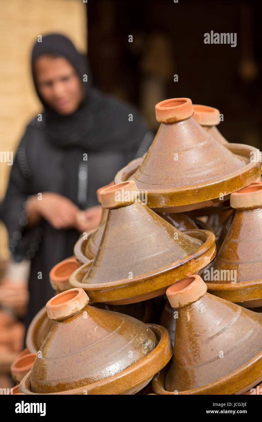 Selezione di marrone tajines marocchine (tradizionali piatti casseruola) impilate sul mercato con la forma di un sfocata donna islamica in background Foto Stock