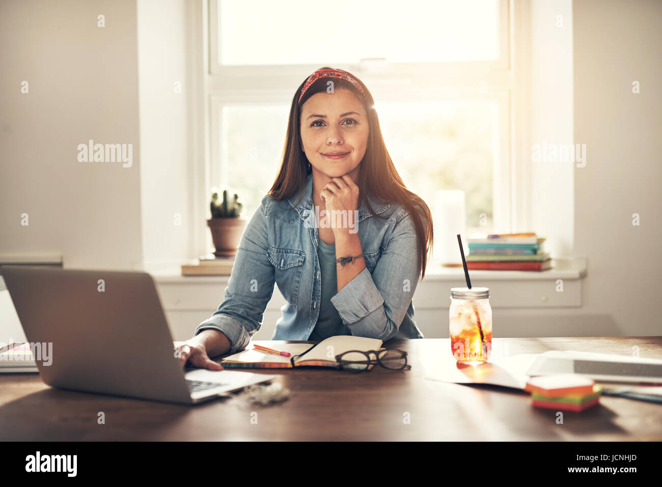 Piuttosto giovane imprenditrice guardando la telecamera seduti al computer portatile in ufficio e sorridente. Foto Stock