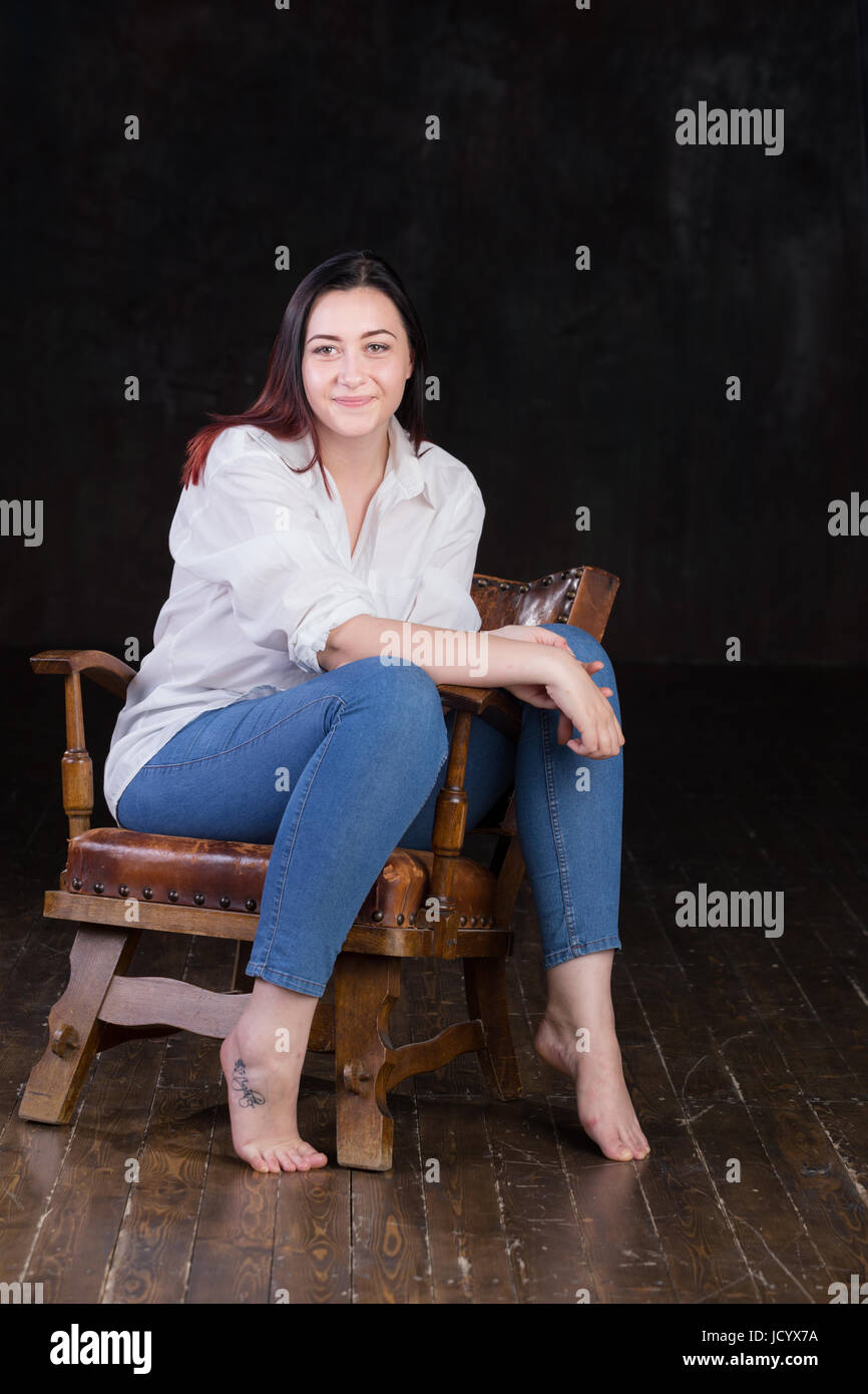 Bella bianca dai capelli marrone femmina con colorati in rosso termina, indossando uomini camicia bianca e jeans blu, seduto in una sedia vintage su sfondo scuro Foto Stock